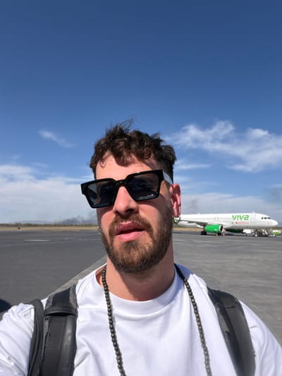 A male subject is depicted in a selfie taken on an airport tarmac at Monterrey International Airport in Apodaca, Mexico. The subject, visible from the chest up, has curly brown hair, a beard, and a mustache, and wears black square-framed sunglasses. He has a nose stud in his left nostril and two silver hoop earrings in his left ear. He is wearing a white t-shirt and a dark beaded necklace, with a black backpack strap visible on his right shoulder. The background features a clear blue sky with sparse white clouds. To the subject's right, a white and green Viva Aerobus aircraft is parked on the tarmac. To the subject's left, beyond the tarmac and a flat, arid landscape, a dark plume of smoke rises in the distance.