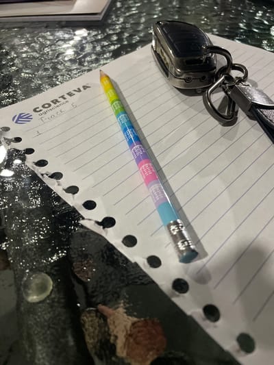 A close-up shot shows a notebook with a Corteva Agriscience logo and "Track 5" written on it. A colorful pencil with multiplication facts printed on it lies across the notebook. A car key fob is also visible, resting on the notebook's pages. The notebook and key fob are placed on a reflective, textured surface that appears to be a table. The background is dark and out of focus, with hints of water droplets or a wet surface. The lighting suggests an indoor setting, possibly in the evening. The image is composed from a high angle, looking down at the objects. The exact location in Slobozia-Dușca, Moldova, is not discernible from the visual information provided.