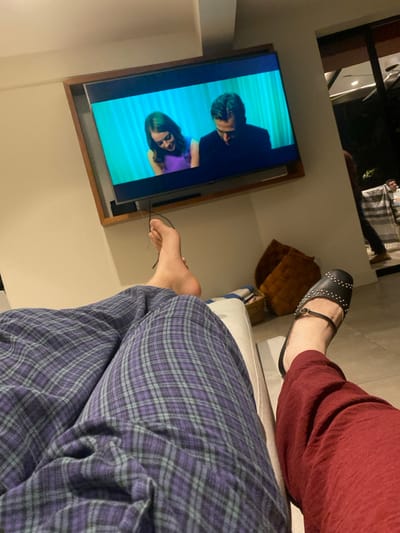 A low-angle shot depicts two people relaxing, likely indoors. One person wears burgundy pants and black, studded sandals. The other is in blue and purple plaid pajamas. They appear to be sitting on a white couch. A television mounted on the wall shows a scene with a man and a woman. Glasses lay next to the leg of the person wearing the pajamas. A brown cushion is visible on the couch behind the person in burgundy.