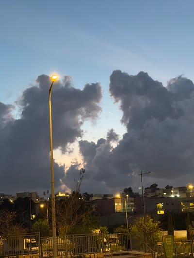 The image presents an urban twilight scene in Harish, Israel, observed from a slightly elevated vantage point. The sky is dominated by extensive, dark gray cumuliform clouds, which are notably parted in the upper center, revealing a segment of lighter blue sky transitioning to a faint orange hue near the horizon. A small, indistinct crescent moon is visible within the cleared sky segment.
A prominent, tall street lamp with a bright yellow pole and an active, illuminated luminaire stands in the left foreground. Below the horizon line, a suburban residential area is visible, characterized by multiple buildings constructed on a gradual slope. Several windows within these structures are illuminated, indicating internal activity. Additional, less prominent street lamps with active lights are distributed throughout the midground, contributing to the ambient illumination.
In the immediate foreground, a dark-colored metal railing or fence is present, bordering an area of sparse vegetation, including several leafless trees or shrubs and some greener foliage. No human figures or direct interactions are visible within the frame. The overall environment depicts a developed residential area at dusk or early night, under significant cloud cover.