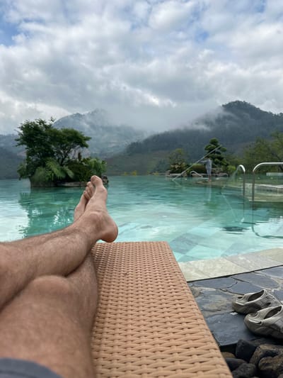 An overhead perspective captures a person's legs and feet resting on a lounge chair, overlooking an infinity pool and a mountainous landscape in Púng Luông, Vietnam.
In the immediate foreground, a small section of dark grey fabric, likely shorts, is visible at the bottom left. Above this, a pair of bare, hairy legs are crossed at the ankles, resting on a light brown, woven-textured chaise lounge chair that occupies the lower right quadrant of the image. The feet are positioned towards the left. To the right of the lounge chair, on a dark, irregular stone surface, a pair of light grey "Keen" brand sandals are placed. Below the sandals, several dark, smooth river rocks are visible.
The midground features a turquoise-blue infinity pool with visible patterned tiling on its floor. The pool water reflects the surrounding environment. Within the pool, on the left side, a natural-looking island densely covered with green trees and ferns is present. Towards the right side of the pool, another smaller, manicured island features a singular, sculptural tree. A person, dressed in light-colored attire, stands in the pool between the right island and the pool edge, holding a long, light-colored pole, possibly for maintenance. Chrome-colored handrails are visible at the pool's edge on the far right.
The background consists of rolling green mountains, partially enveloped in mist and low-hanging grey and white clouds. Patches of lighter blue sky are discernible through the cloud cover. The overall atmospheric condition appears overcast.