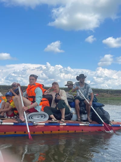 A group of people are on a raft on a body of water under a blue sky with white clouds. The raft is red with yellow and blue accents. Several people are wearing orange life jackets. One person in the foreground on the left is wearing a blue shirt, red shorts, and an orange life jacket, and is holding a paddle. Another person in the foreground on the right is wearing a camouflage shirt and a black cowboy hat, and is also holding a paddle. There are several bags and rolled-up items on the raft. The background shows a grassy bank with some vegetation. The scene appears to be a recreational activity on a river or lake.