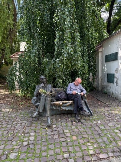 The scene is set in a small courtyard in Kraków, Poland, at the coordinates 50.053, 19.948. A man is sitting on a stone bench, reading a book. The bench is positioned under a large weeping willow tree, providing shade. Behind the bench is a white wall, adorned with plaques. The ground surrounding the bench is covered in cobblestones. The man appears to be middle-aged and is dressed in a light-colored button-down shirt and dark-colored pants. He is wearing glasses. The bench is adorned with a bronze sculpture of a man, also reading a book. The sculpture is made of bronze and appears to be a depiction of a prominent figure. The man and the sculpture both appear to be absorbed in their reading.