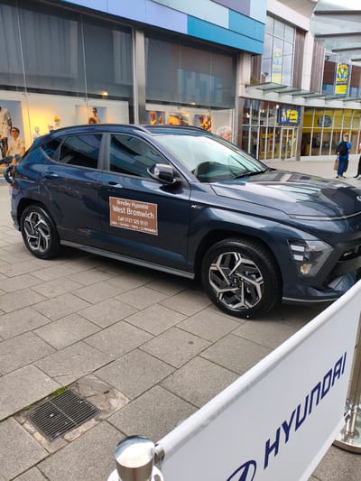 A dark blue Hyundai Kona N Line is parked on a paved pedestrian area in front of a row of shopfronts. A large white banner with a blue Hyundai logo and text is partially visible in the foreground. The car has a sticker on its driver's side door that reads "Brindley Hyundai West Bromwich" and provides a contact phone number and service details. Behind the car, shop windows display mannequins and merchandise. In the background, other shops with signage, including "Monkey Monkeys" play centre, are visible. Several people are walking in the background, including a person with a blue backpack. The environment appears to be a public walkway or town centre, likely in Sandwell, United Kingdom, given the mention of West Bromwich on the car's sticker.