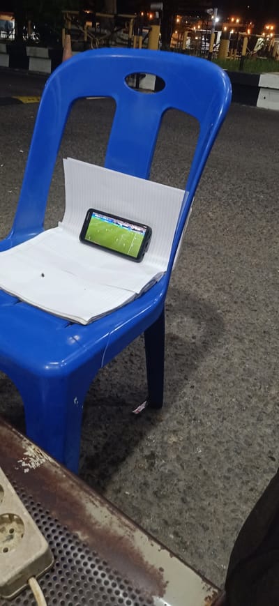 A bright blue plastic chair sits on an asphalt surface, likely outdoors as suggested by the dark, ambient lighting of what appears to be evening or night. The chair is occupied by a stack of white paper, with a smartphone placed on top. The phone's screen displays a soccer game in progress, with players visible on the green field. The background is blurred, but hints of outdoor lighting, possibly from street lamps or buildings, are visible, along with some greenery and what looks like a fence or barrier. To the lower left of the image, a section of a rusty metal object with a power strip attached is visible, further suggesting an outdoor setting perhaps at a casual gathering place or a street-side stall. The overall mood is relaxed and informal, with the focus on the soccer match being viewed on the phone. There is no visible text in the image.