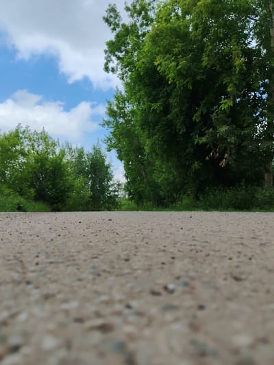 The image captures a low-angle view of an asphalt path or road, with lush green trees lining both sides and extending into the background. The sky above is blue with scattered white clouds. The scene appears to be an outdoor, natural setting within the city of Сокол, Russia, possibly a park, rural road, or a walking trail. There are no people or animals visible in the frame. The focus is primarily on the textured surface of the path in the foreground, with the foliage and sky serving as a backdrop.