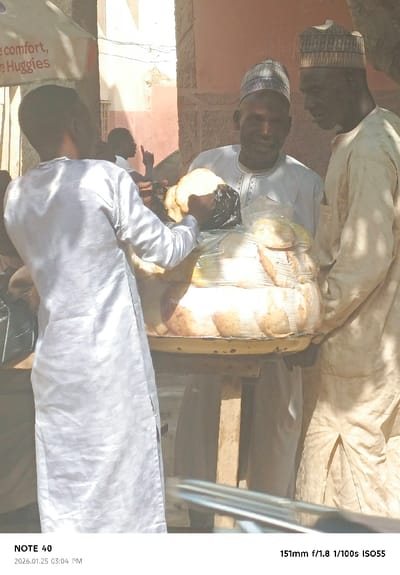 The image depicts an outdoor market scene in Tudun Wazurchi, Nigeria, during daylight hours. Three adult males are visible in the foreground and midground, with a fourth individual partially visible in the background. In the center, a large, circular, yellow metal tray filled with numerous light-brown baked goods, likely bread rolls, is supported by a wooden stand. Many of the baked goods are individually wrapped in clear plastic, and a black plastic bag is positioned among them.

A male figure with his back to the camera, wearing a light-colored, long-sleeved tunic, stands on the left, reaching towards the tray. Directly behind the tray, a male figure wearing a white tunic and a patterned cap smiles towards the scene in front of him. To the right, another male figure, wearing a light-colored tunic and a similar patterned cap, stands in profile, observing the interaction. In the blurred background, a person is seen gesturing upwards with their right hand. Overhead, a light-colored awning or umbrella is partially visible, displaying red text that includes "comfort," and "Huggies." The background features reddish-brown building walls under bright sunlight.