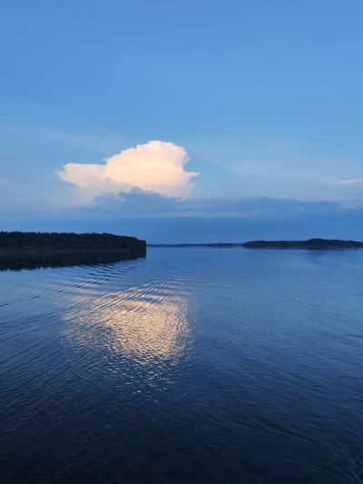 The image depicts a serene natural landscape with a vast body of water under a twilight sky. The water surface is calm, reflecting the soft hues of the sky and a large, luminous cloud formation. Gentle ripples emanate from the foreground, suggesting movement, possibly from a boat or wind.
On the left side of the frame, a densely forested landmass stretches along the water's edge, its dark silhouette contrasting with the brighter sky and water. The trees appear to be coniferous, forming a thick, continuous line. In the distance, a similar, but more elongated, landmass is visible on the horizon, also covered in trees. The sky above is a gradient of deep blue at the bottom, transitioning to a lighter, clearer blue towards the top. A prominent cumulus cloud, illuminated from within by warm, golden light, is the focal point of the sky. Other scattered clouds in softer shades of blue and grey are also present.
The scene suggests a location in a natural setting, likely a lake or a wide river, given the expanse of water and the presence of forested shores. The lighting indicates either early morning or late evening, characteristic of twilight. The overall atmosphere is peaceful and tranquil.
Given the provided context, this scene could be located near Ul'yanovo, Russia, a region known for its numerous lakes and forested areas, particularly within the European part of Russia. The image captures a typical vista of the Russian countryside during the summer or early autumn twilight hours.