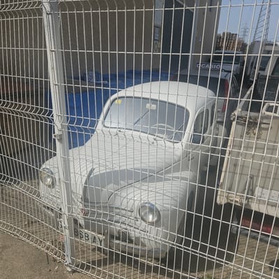 The image shows a white vintage car parked behind a metal fence. The car appears to be in a lot or yard, with other vehicles and stacks of pallets visible in the background. A sign that says "OCASIO" is visible on the back of a silver car behind the vintage car. The ground is paved with gravel and dirt. The scene is illuminated by sunlight, casting shadows from the fence onto the car. The overall impression is one of an outdoor storage area or a used car lot. The location in El Perelló, Spain, suggests a Mediterranean coastal town.