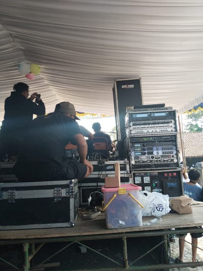 The image depicts an outdoor gathering under a white tent, likely in Ngadirejo, Indonesia, given the context. The scene focuses on a sound system setup, with various audio equipment stacked on racks and cases. Several individuals are present, some appearing to be involved with the setup or attending the event.
In the foreground, a person in a black t-shirt and shorts is seated on a black flight case, facing away from the camera, engrossed in their surroundings. To the left, another person in a black jacket is holding a smartphone, possibly taking a photo or video. In the background, other individuals are visible, including a person wearing a cap and a person in a blue t-shirt.
The environment includes a white tent covering the area, providing shade. Various pieces of audio equipment, including speakers, amplifiers, and mixers, are arranged on a wooden platform. Several black flight cases are also present, suggesting portability and protection for the equipment. A pink and yellow bucket, a white plastic bag, and a small cardboard box are scattered on the platform. In the upper left corner, three balloons (white, light green, and pink) are suspended from the tent. The background shows glimpses of tiled roofs and greenery, indicative of a semi-rural or residential setting. The overall impression is that of a sound system setup for an outdoor event or performance.