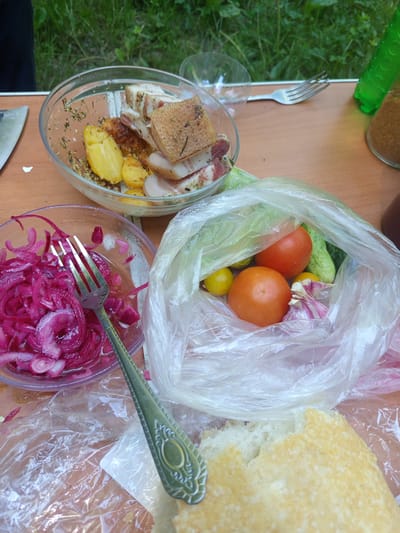 The image depicts a close-up, overhead view of a wooden table set with food items, likely outdoors due to the visible greenery in the background and the casual arrangement of the meal. In the foreground, a torn piece of bread rests on a clear plastic bag. To the left, a glass bowl contains thinly sliced red onions, with a silver fork resting inside. Behind this bowl, another glass bowl holds pieces of cooked potatoes and cured meat with herbs. Further back, a clear plastic bag is filled with fresh vegetables including tomatoes, cucumbers, and garlic. A green bottle and a glass are partially visible in the upper right of the frame. The overall scene suggests a picnic or an outdoor meal. The location is identified as Giaginskaya, Russia.