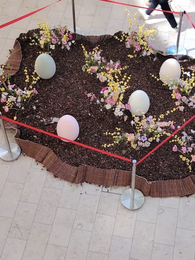A high-angle shot depicts an Easter display in Kehl, Germany, set on a tiled floor. The display consists of a bed of dark mulch enclosed by a border of woven reeds. Within the mulch are four large decorative eggs: one light green, one pink, and two white. The eggs are surrounded by an assortment of pink and yellow flowers. The display is cordoned off by a red rope attached to silver stanchions. In the upper right corner of the frame, the legs of a person walking by are visible.