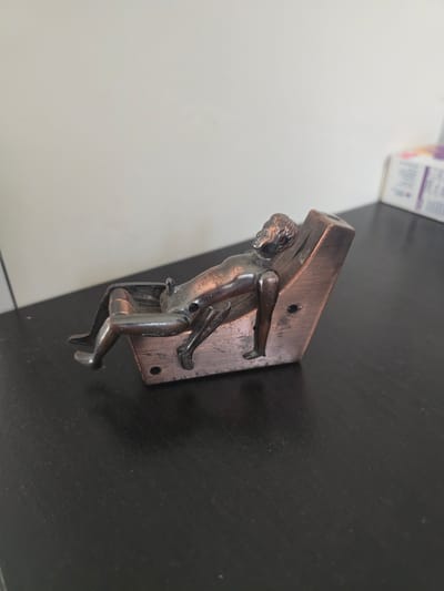 The image shows a small, metallic figurine of a person reclining on a lounge chair. The figurine appears to be made of a bronze-colored metal and has a distressed, aged patina. It is positioned on a dark, flat surface, likely a table or desk. The background is a plain, light-colored wall. The figurine is detailed, with clear representations of the human form, including facial features, limbs, and clothing. The chair is also depicted with some detail, suggesting a reclining design. The overall impression is of a decorative or novelty item. The location in Bönningstedt, Germany is not ascertainable from the image.
