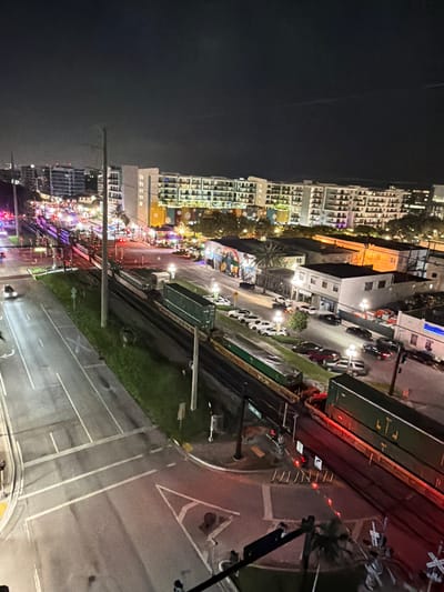 This is an aerial, nighttime photograph of a city scene in Hollywood, Florida, USA. In the foreground, a train with green cargo containers is stopped on the tracks. The train crosses a road where traffic lights are visible, and other vehicles are stopped or moving. A street with parked cars and buildings lines the opposite side of the train tracks. Further in the background, a row of multi-story buildings, likely apartment complexes, are illuminated, with some displaying colorful murals on their lower levels. The sky is dark and cloudy, with a faint light suggesting urban illumination.
