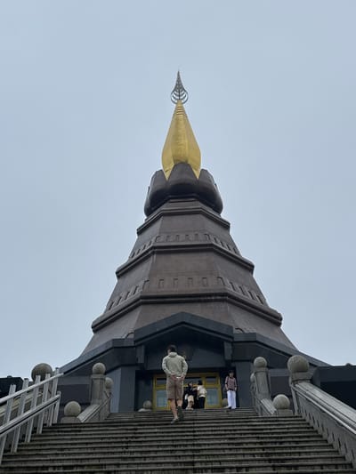 This image depicts the Great Noble Relics Pagoda at Doi Inthanon National Park, located near the city of Chang Khoeng, Thailand. The pagoda, a towering structure with a dark, textured base and a prominent golden spire, dominates the frame against a pale, overcast sky. A wide, stone staircase leads up to the pagoda's entrance, where several individuals are ascending. On the left side of the stairs, a man in a light green jacket and khaki shorts walks away from the camera. Further up the stairs, a group of people, including a woman in white pants and a dark top, and others in casual attire, are visible near the entrance. The staircase is flanked by ornate stone balustrades topped with spherical finials. The environment appears to be a tourist destination, with visitors engaging in the act of climbing the stairs to view the monument.
