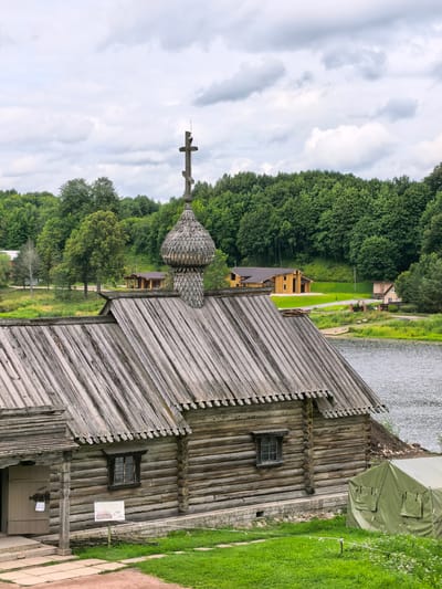The image depicts a historic wooden church with a prominent onion dome topped with a cross, situated in Staraya Ladoga, Russia. The church is constructed from large, rough-hewn logs, with a weathered wooden roof. Small, multi-paned windows are visible on the church's facade. In the foreground, a green tent is pitched on a grassy area. Behind the church, a body of water, likely a river, stretches into the distance, bordered by lush green trees and rolling hills. Several other wooden buildings, appearing to be houses or small structures, are scattered along the shoreline and further inland. The sky is overcast with dramatic clouds. The scene conveys a sense of historical preservation and natural beauty.