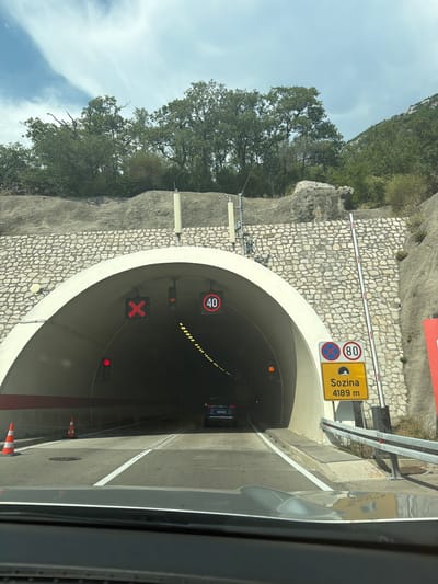 This is a media file of a road tunnel entrance. The tunnel is illuminated with yellow lights, and a car is visible inside. Traffic signs indicate a speed limit of 80 km/h and provide information about the tunnel's name, "Sozina," and its length, 4189 meters. Additionally, a red "X" symbol is displayed, suggesting a closure or restriction.
The scene appears to be set in daylight with a clear sky visible above the tunnel entrance. The tunnel is surrounded by a rocky hillside covered with trees. The overall impression is one of travel and infrastructure, with the presence of traffic control devices and a clear indication of the tunnel's function and name. The location is identified as Đurmani, Montenegro. The image is taken from inside a vehicle, looking towards the tunnel entrance.