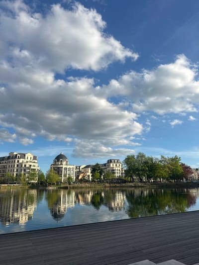 The image captures a serene waterscape under a bright blue sky dotted with scattered, fluffy white clouds, indicating clear and fair weather during the daytime. The scene is viewed from a dark wooden planked deck or walkway that extends along the right side of the foreground, bordering a calm body of water, likely a canal or lake.
The water acts as a mirror, creating clear reflections of the sky, clouds, and the buildings and trees lining the opposite bank. Along this far bank, a series of elegant multi-story buildings with light-colored facades and dark roofs are visible. A particularly notable building features a distinct dark, domed roof. Interspersed among the architecture are lush green trees, with some displaying hints of reddish foliage.
No people or specific activities are visibly taking place, contributing to the tranquil and peaceful atmosphere of the urban setting. The architecture suggests a European location, which aligns with the provided context of Clamart, France. There is no visible text in the image.