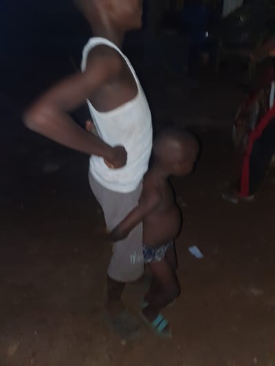 The image captures an intimate moment between an adult and a child in a dimly lit, possibly nighttime or indoor setting. The photograph is taken from a high angle, looking down.
The primary subjects are two individuals, both appearing to have dark skin tones. On the left, an adult is partially visible, wearing a white tank top. A young child, clad in patterned shorts and light blue or green sandals, is lying horizontally across the adult's stomach or lap. The child's head is turned away from the camera, and their body appears relaxed, suggesting comfort or rest.
The background is predominantly dark, indicating poor lighting. The floor appears to be made of dirt or dark earth, suggesting a rustic or informal environment. Several small, light-colored objects, possibly debris or paper, are scattered on the ground near the child's feet. In the upper part of the image, a blurry red and black object is faintly visible.
The overall scene conveys a quiet and possibly tender interaction between the two individuals. The low light conditions contribute to a sense of privacy or late hours. No discernible text is visible in the image. Given the context of Idah, Nigeria, the setting's simple nature and dim lighting could be characteristic of some local environments.