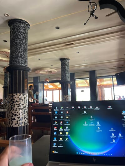 A person's hand holds a glass of pale green liquid in the foreground. To the right, a laptop screen displays a Windows desktop with numerous icons and a colorful background. In the background, a restaurant or cafe setting is visible with dark pillars adorned with black and white speckled patterns. A man in a patterned shirt is seen walking in the middle ground. The overall ambiance suggests an indoor dining or working environment with natural light filtering in from large windows in the rear. The location is specified as El Hajeb, Morocco.