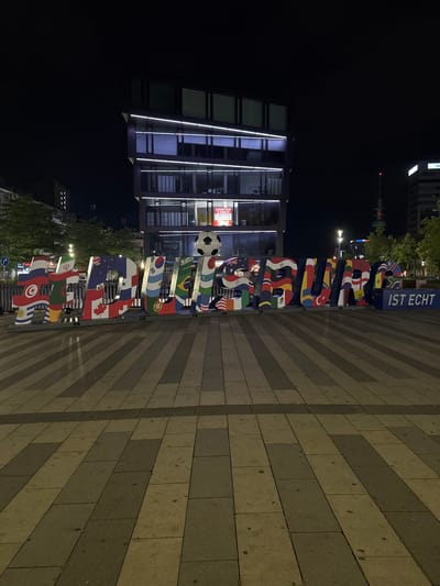 The image shows a large art installation in the city of Duisburg, Germany, at night. The installation consists of three-dimensional letters spelling out "FUSSBALL IST ECHT" (Football is Real). Each letter is decorated with the flag of a different country, representing nations that participate in football. A soccer ball is placed atop the letter "A" in "FUSSBALL". The installation is situated in a public square paved with interlocking rectangular stones, some of which are illuminated by streetlights. In the background, modern buildings with illuminated windows and facades are visible, along with trees and a telecommunications tower. The overall scene is well-lit, suggesting a public space designed for events or as a tourist attraction. No people are visible in the image.