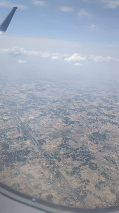 An aerial view from an airplane window shows a vast expanse of rural landscape with fields, scattered vegetation, and buildings. The fields are irregular in shape and appear to be harvested or fallow, with varying shades of brown and beige. Darker green patches indicate clusters of trees or densely vegetated areas. A road or highway runs diagonally through the scene, bordered by more settlements and fields. Small villages or hamlets are visible, characterized by clusters of buildings and trees. In the upper left quadrant, a portion of the airplane's wing is visible against a bright blue sky with scattered white clouds. The overall impression is of an agricultural region from a high vantage point. The specific location in Babatpur, India is characterized by this agricultural pattern, with the highway potentially being a major artery connecting to urban centers. The environment appears dry and possibly during a harvest or post-harvest season.