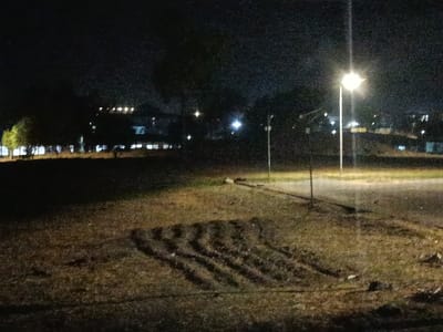 The image displays a low-light night scene in Idon Kasa, Nigeria. The foreground and mid-ground consist of an unpaved, uneven area of dark earth and sparse vegetation, possibly grass or weeds, appearing brown and dark green under ambient illumination. Distinct parallel tire tracks are visible in the soil towards the bottom-center. To the right, a tall, slender pole supports a brightly illuminated streetlamp, which is the primary light source in the scene, casting a yellow-white glow. A second, darker, and slightly shorter pole stands further to the left of the prominent streetlamp. A dark, linear strip runs horizontally across the mid-ground, potentially indicating a road or path. In the background, dark silhouettes of trees are visible, behind which a cluster of illuminated windows and lights from multiple structures or buildings can be observed against the dark night sky. No individuals, vehicles, or active subjects are present.