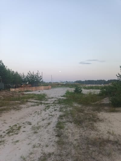 The image depicts an outdoor scene in Yasnyska, Ukraine. The foreground is a dirt path with sparse vegetation. To the left, a pile of wooden pallets is stacked, and a wooden fence runs along the edge of the path. Beyond the fence, a line of trees is visible. In the distance, a pale moon is present in the sky, and a faint, wispy cloud can be seen. The overall environment appears to be rural or semi-rural, possibly a construction site or an area undergoing development, given the presence of building materials. The sky suggests it is either early morning or late evening due to its pale hue and the presence of the moon.