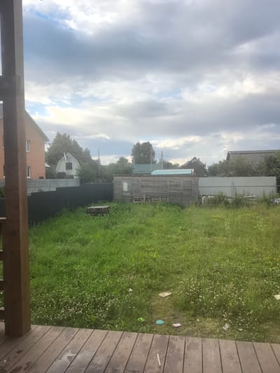 The image depicts an outdoor scene in Pogorelki, Russia, on a cloudy day. In the foreground, a wooden porch with weathered planks is visible, with water stains indicating recent moisture. Beyond the porch, a grassy yard stretches out, dotted with small white and pink flowers, along with some scattered debris including a blue plastic item and some paper waste. A dark green fence runs along the edge of the yard, separating it from what appears to be a more developed area.
In the middle ground, a long, narrow wooden shed with a window is situated. To the left of the shed, a wooden pallet is placed on the grass. Behind the fence, several buildings are visible. On the far left, a portion of a multi-story brick building with white-trimmed windows can be seen. Next to it, a white house with a distinctive arched roofline and a dark-framed window is present, surrounded by trees. Further to the right, other structures with pitched roofs, some covered in dark tiles, are partially obscured by trees and foliage. A light-colored corrugated metal fence runs behind the wooden shed. The sky above is filled with heavy, grey clouds, with breaks of lighter sky and sunlight peeking through in some areas. The overall impression is that of a rural or semi-rural residential area.