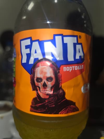 A plastic bottle of Fanta, positioned horizontally, is the main subject of the image. The bottle contains a bright yellow-orange beverage, consistent with an orange-flavored drink. Its label is predominantly orange, featuring the Fanta logo in large, stylized blue and white letters.
A notable detail on the label is a prominent illustration of a skull character, which has a reddish-orange hue and is depicted wearing a dark, hooded garment, resembling the iconic imagery often associated with the band Ghost or similar dark rock aesthetics. To the left of the skull and above the Fanta logo, a red banner contains white Cyrillic text that reads "портокал" (portokal), which translates to "orange," confirming the flavor and suggesting a Bulgarian context, consistent with the provided location context of Kraishte, Bulgaria.
Other visible text includes a small white circle on the upper right of the label showing "100 мл" (100 ml) and "ккал" (kcal), indicating nutritional information per 100 milliliters. A registered trademark symbol (®) is also visible near the top right of the Fanta logo.
The bottle rests on a dark, slightly reflective surface. Bright light reflections are visible on the bottle's surface, particularly on the top left, indicating artificial lighting in an indoor setting. No specific activity or event is taking place; it is a still shot of the product.