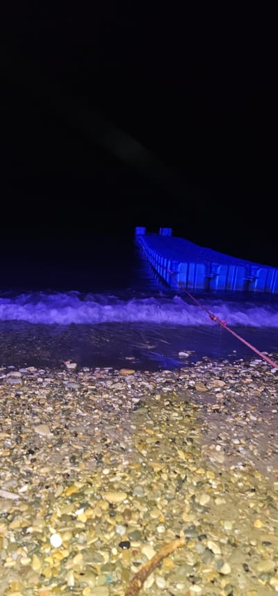A vertical, medium-shot, nighttime photograph captures a beach in Lermontovo, Russia. In the foreground, a shore of mixed-color pebbles is wet, reflecting the purple light of the waves. The water, illuminated by a blue, artificial light source, crashes onto the shore in small, foamy waves. In the mid-ground, a blue, modular pontoon dock extends into the dark water, with a reddish-brown rope tied to its side and leading towards the shore. The sky is completely black, indicating the absence of moonlight and a lack of ambient light.