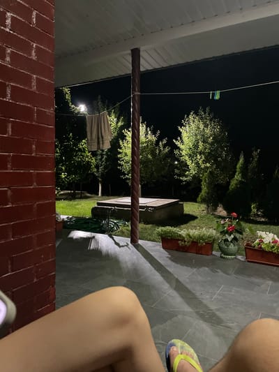 The image captures a dimly lit outdoor scene at night, likely in a residential backyard in Slobozia-Dușca, Moldova. The foreground is dominated by a section of a red brick wall. A person's legs and feet, wearing a yellow and blue flip-flop, are visible on a dark gray tiled patio, suggesting a casual, relaxed atmosphere.

The middle ground reveals a well-maintained garden with lush green grass and various plants. A few potted plants, one with white and red flowers, are placed on the patio. A coiled green garden hose rests on the grass near a small, light-colored structure. Trees with illuminated foliage form a backdrop, with a single outdoor light fixture visible, casting a warm glow and creating shadows on the ground. The overall impression is one of a peaceful evening outdoors, possibly after dusk. The scene appears dry, with no indication of rain. There is no visible text in the image.
