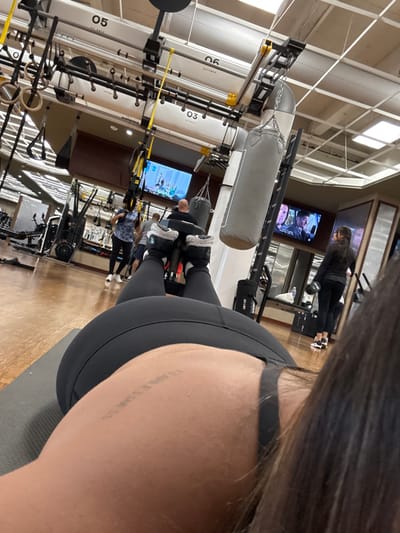 The image depicts the interior of a fitness center, specifically a Life Time facility located at 1000 Airport Rd, Plainsboro Township, New Jersey, United States.

The scene is viewed from a first-person perspective, looking forward over the back of a person lying prone on a dark gray exercise mat. This individual wears black athletic leggings and a black sports bra. A tattoo reading "FEARLESS" is visible on their upper left back. Their feet are elevated, shod in black and white athletic shoes, with dark ankle weights or resistance bands visible.

In the midground and background, several individuals are present. A bald male in a dark shirt is partially visible, seated or resting. A female with dark hair, dressed in black athletic wear, stands near a bench, holding a black object in her left hand. Another female, partially obscured, wears a patterned top and dark leggings.

The environment features a large open space with a light brown floor and a white ceiling equipped with integrated lighting, ventilation, and an extensive black and yellow overhead metal frame from which multiple gray cylindrical punching bags and yellow TRX suspension straps are suspended. Numbered stations ("03", "05", "06") are marked on the overhead structure. Two large flat-screen televisions are mounted on a far wall; the left screen displays a fitness class, and the right screen shows a scene from a television program or movie. Various fitness machines and equipment are distributed throughout the facility.