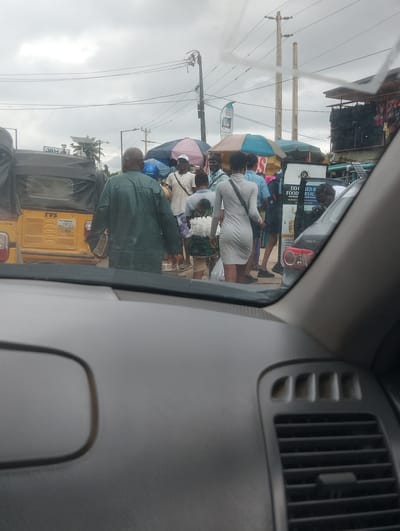 The image depicts a street scene in Iju, Nigeria, on an overcast day. In the foreground, the interior of a car is visible, with the dashboard and windshield framing the view. The scene outside the car includes a yellow auto-rickshaw with the TVS logo and a license plate that reads "JJJ-62LDP". Several people are gathered around the auto-rickshaw and surrounding stalls, some under colorful umbrellas. A man in a green traditional outfit is prominent, facing away from the camera. A woman in a light grey dress walks away from the auto-rickshaw, carrying a bundle. Other individuals, some appearing to be vendors or shoppers, are also present. Various stalls with umbrellas and goods are set up along the street, suggesting a market or roadside commerce. Power lines and utility poles are visible in the background, characteristic of an urban or semi-urban environment. A dark-colored car, possibly a Toyota, is parked on the right side of the image.