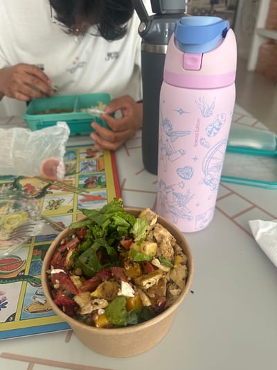 This image captures an overhead view of a meal setting indoors, likely at a café or home in Sanctorum, Mexico, suggested by the Loteria-style placemat.

In the foreground, a person with dark hair and glasses, wearing a white t-shirt, is visible from the chest up, with their head mostly obscured. Their hands are seen interacting with a teal bento box, possibly arranging or selecting food from its compartments.

To the right, a brown paper bowl contains a colorful salad, featuring green lettuce, diced pieces of what appears to be chicken or tofu, bright red strawberries, yellow mango or peach pieces, white crumbly cheese (possibly feta), and black sesame seeds. Next to the bento box is a clear plastic bag holding a small orange container, likely for dressing, and a clear plastic fork.

Dominating the left side of the frame is a pink water bottle with a blue and light pink lid. The bottle is adorned with various light blue illustrations including angels/cherubs, birds, stars, dice, and a seashell-like spiral. The text "Special Edition" is clearly visible on the bottle. A dark gray thermal bottle is partially visible beneath the pink one.

The items rest on a white table with subtle pink-lined tiles. Beneath the salad bowl and near the person's hands is a colorful mat or placemat depicting various illustrated cards, characteristic of the traditional Mexican Loteria game, with numbers like "24" and "3" visible on some cards. A white napkin is also seen in the upper right. The scene appears to be taking place during the daytime.