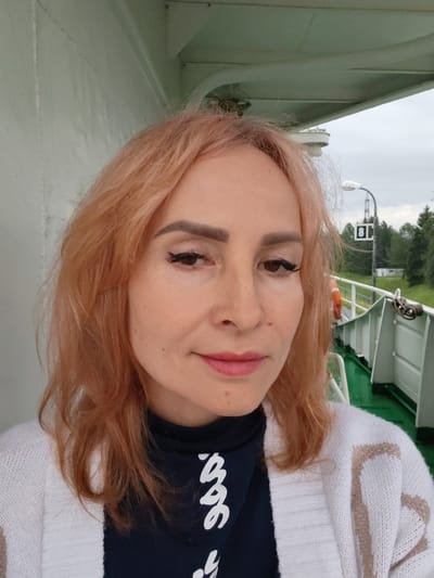 A woman with shoulder-length, light auburn hair, wearing a dark blue shirt with white cursive writing and a white knitted cardigan with light brown stripes, poses for a selfie outdoors. She has on light makeup, including eyeliner and lipstick. The background appears to be the deck of a boat or ship, with a green deck and white railing. Beyond the railing, there are trees and a cloudy sky. A lamppost with a sign that has the number 8 on it is visible in the background, as is an orange life preserver. The setting appears to be a waterway, possibly a canal or river, in or near the city of Belousovo, Russia.