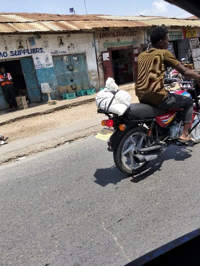 A daytime street scene in Mkomani ward, Kenya, depicts a commercial thoroughfare under a clear, light blue sky. In the mid-ground, a dark-skinned male in a brown striped shirt, dark trousers, and sandals is positioned on a red "BOXER 150" motorcycle, with a large white sack secured to the rear luggage rack by black straps. The motorcycle displays Kenyan license plate KMCX 460B. The paved asphalt road in the foreground exhibits minor surface cracks, and the motorcycle casts a dark shadow. To the left, an unpaved dirt shoulder separates the road from a row of single-story commercial buildings in the background. These structures feature rusty corrugated metal roofs and weathered white-painted facades with peeling paint. The leftmost building, partially open, shows signage "NO SUPPLIERS." and "FUNDI SAA"; a dark-skinned male wearing an orange high-visibility vest stands in its doorway. Adjacent to it, a shop with closed blue metal doors is visible. To the right, another establishment with a red entrance bears the green text "IMPERIALIST ENTERTAINMENT," with a darkened interior and an indistinct figure within. A small purple flag is visible on the roofline.