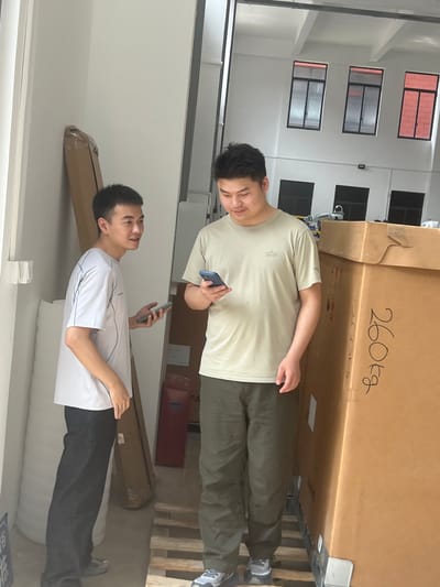 Two adult males are standing near a large cardboard box labeled "260Kg" in a warehouse setting. The man on the left, wearing a white t-shirt and black pants, is holding a mobile phone and looking towards the man on the right. The man on the right, wearing a light brown t-shirt and green pants, is looking down at his mobile phone. Behind them, there are several other cardboard boxes, some stacked, and industrial shelving. The environment appears to be an indoor storage or distribution facility, potentially in Shuangfu, China, given the context. The lighting suggests it is daytime, with natural light coming from an opening on the left side of the frame.