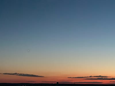 A wide shot captures the vast expanse of the sky during dusk. The sky transitions from a deep blue at the top to a soft orange and pink hue near the horizon, indicating the time of day is sunset. A single bright star or planet is visible in the upper left quadrant of the image. Wispy clouds are scattered across the lower portion of the sky, some tinged with the colors of the sunset. Below the sky, a dark silhouette of land is visible, suggesting a landscape or cityscape. A few distant lights are faintly visible on the horizon, hinting at human settlement. The overall mood of the image is serene and peaceful, evoking the beauty of the natural world during twilight. There is no visible text in the image.