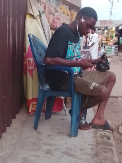 A young Black male wearing a black t-shirt with a green and blue design and tan shorts is seated on a blue plastic chair. He is holding a black plastic bag in his right hand and looking down at a mobile device in his left hand. He has white earbuds in his ears. The background shows a market stall with shelves stocked with various goods. There are other people in the background, including one person wearing a white robe. The ground is paved with concrete and dirt. The scene appears to be outdoors. There is no indication in the image that this scene is located in Birmingham, United Kingdom.