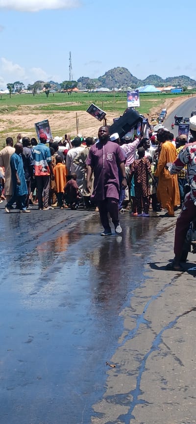 The image depicts a street scene in Kayarda, Nigeria, on a sunny day with a clear blue sky. The foreground shows a wet asphalt road, reflecting the sky and the people. A diverse group of people, predominantly men, are gathered on the road and the adjacent sandy shoulder. They are dressed in various traditional African attire, including kaftans, agbadas, and wrappers. Some individuals are holding up signs or posters, suggesting a gathering or procession. In the background, a road stretches into the distance, with vehicles and a telecommunications tower visible. The landscape features rolling hills and scattered buildings, characteristic of a rural or semi-urban setting in Nigeria.