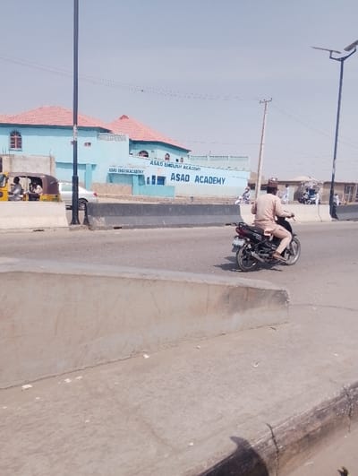 This image depicts a daytime outdoor scene in Chiranchi, Nigeria, featuring a multi-lane asphalt road with a concrete median barrier. In the midground, an individual dressed in light-colored traditional Nigerian attire is operating a black motorcycle, traveling from right to left across the frame. In the left background, a light blue building with a reddish-orange tiled roof is prominently displayed, bearing the signage "ASAD ENGLISH ACADEMY" and "ASAD ACADEMY." Adjacent to this building, a yellow motorized tricycle, occupied by at least three individuals, is visible. A tall, black utility pole stands in the left midground, with power lines extending overhead across the clear sky. In the right background, another black pole with a solar panel, characteristic of a street light, is present. Additional, less distinct buildings and several other individuals, some near parked motorcycles, are visible further down the road on the right. The foreground consists of a concrete curb and a sloping concrete structure.