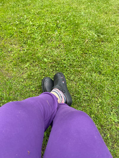 The image shows a person's legs, clad in bright purple sweatpants, resting on a patch of green grass. The person is wearing black Crocs and striped socks. The grass appears to be a lawn, possibly in a park or backyard, with some clover mixed in. There are no city landmarks or identifiable structures visible in the image, so it is impossible to determine the specific location in Ленинградская, Russia, or any other city. The scene is static and focuses on the lower half of the person's body and the surrounding ground.