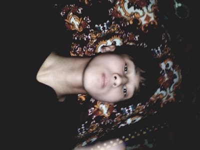 One individual, a young person with dark hair and light skin, is depicted lying down. The person's head is slightly turned, and their eyes are directed towards the camera. They are wearing a dark-colored garment that covers the upper torso and left shoulder. The individual is positioned on a surface covered by a patterned fabric, which features an ornate, repeating design in dark, orange, red, yellow, white, and light blue hues. The environment is dimly lit, casting a subdued tone over the scene. The scene takes place in the city of Bagat, Uzbekistan.