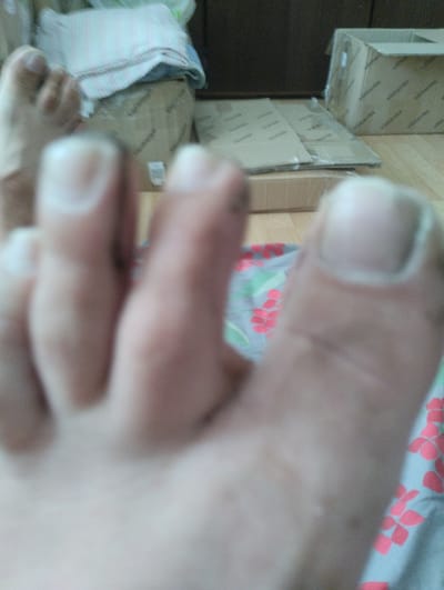 The image is a close-up, out-of-focus shot of a person's toes. The toes are light-skinned and appear to be resting on a light blue fabric with a pattern of pink flowers. In the background, several cardboard boxes are visible. One box is open and appears to have some fabric or bedding inside. Another box is stacked on top of it. Further in the background, another larger cardboard box is visible, and to the left of that, a dark wooden surface is present, possibly a cabinet or dresser. The overall lighting suggests an indoor setting, possibly during the daytime given the ambient light. There is no clear indication of the specific time of day, weather, or emotions. No visible text can be clearly discerned in the image. The setting appears to be a room where items are being stored or unpacked, given the presence of numerous boxes.