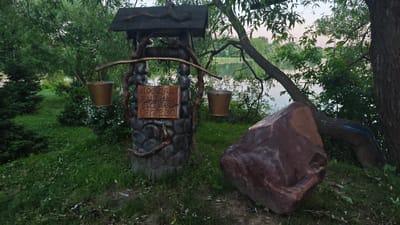 In the village of Shchapovo, Russia, an old-fashioned well stands beside a lake. The well, made of stone and wood, has a roof and two buckets hanging from its sides. A wooden sign on the well reads "Прошло жизнь полей дерево", which translates to "The life of the fields has passed, wood". The surrounding area is covered in lush green grass and trees, with the tranquil lake reflecting the sky and foliage. A large, reddish-brown boulder rests to the right of the well, adding a natural, rugged element to the scene. The overall atmosphere is peaceful and serene, evoking a sense of rustic charm and the passage of time.