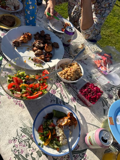 The image captures an al fresco meal, likely a picnic or barbecue, held on a bright, sunny day in Virši, Latvia. A table, covered with a floral-patterned tablecloth, is laden with a variety of dishes, with strong shadows cast across it by the direct sunlight.
At least two individuals are present, indicated by visible arms and hands. One person, wearing a patterned top, is seen on the left side of the frame, holding a fork.
The spread of food includes:
* A large white plate filled with grilled meat pieces, resembling shashlik or kebabs, along with grilled chicken parts.
* Another plate holding a piece of grilled chicken, a creamy potato salad or similar side, and a fresh salad made of chopped tomatoes, cucumbers, peppers, and dill.
* A white bowl containing a rice dish, possibly pilaf, mixed with vegetables.
* A clear plastic container with a vibrant red beetroot salad.
* A large glass bowl filled with a fresh salad of chopped tomatoes, lettuce, dill, and spring onions.
* Dark bread on a yellow plate. Other food items in clear plastic bags and containers are also visible.
Beverages include two cans of Rekorderlig Premium Swedish Cider. One pink can is labeled "Watermelon-Citrus" with "4.5% vol", and a purple can is labeled "Passionfruit". A glass containing an orange liquid, possibly juice, is also present. A blue child's container decorated with white duck illustrations sits on the table. Part of a blue plate shows text that includes "Bianca's Skarptun".
The scene depicts a casual and relaxed outdoor gathering, where people are enjoying food and drinks on a clear, sunny day, suggested by the distinct shadows. The background shows green grass, indicating a garden or park setting.