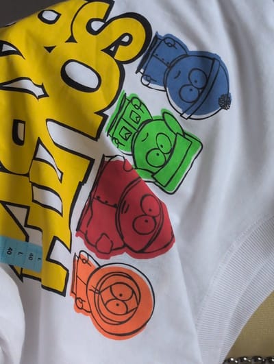 This is a close-up shot of a white t-shirt featuring a colorful graphic of four characters from the animated TV show "South Park." The characters, drawn in a simplified, cartoonish style with black outlines and solid colors, are arranged from left to right: a blue character wearing a blue hat with a pom-pom, a green character wearing a green hood, a red character wearing a red hooded jacket, and an orange character wearing an orange hood. Below the characters, large yellow lettering with black outlines spells out "SOUTH." The t-shirt appears to be made of cotton and has a ribbed neckline. A small blue tag with the letter "L" and the number "40" printed on it is visible near the bottom right of the image. The lighting suggests an indoor setting. The overall impression is casual and playful, typical of merchandise related to the show.