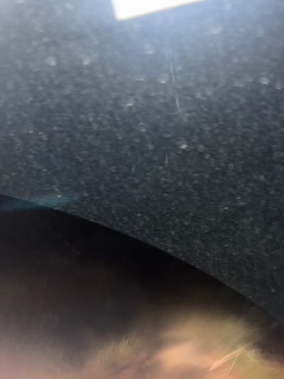The scene is located in La Marquesa, Mexico. The image presents an obscured view, horizontally bisected by a curved line. The upper two-thirds of the frame consist of a dark, textured surface, possibly charcoal gray or black, uniformly covered with numerous small, irregularly shaped, light-colored particles, suggesting dust or debris. A bright, rectangular light source, cropped, is visible at the very top edge. Faint, thin, vertical linear marks are discernible on this upper surface. The lower third of the frame reveals a smooth, dark, curved surface, which reflects a heavily blurred and out-of-focus external scene. This reflection is characterized by diffuse warm tones, including shades of orange, yellow, and reddish hues, along with vague, elongated, lighter patterns. No identifiable people, distinct objects, or specific actions are visible within the frame.