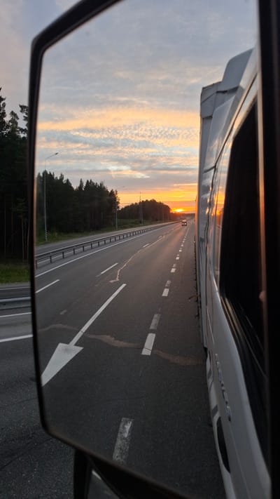 The image, taken from the perspective of a truck driver, shows a sunset view of a highway in Bogandinsky, Russia. The foreground is dominated by a side-view mirror reflecting the highway and the sky. The highway itself appears to be a multi-lane road with white lane markings. On the left side of the road, there is a forest line and a guardrail. In the distance, a lone car is visible on the highway. The sky is painted with hues of orange and yellow from the setting sun, with scattered clouds. The side of a white truck is partially visible on the right side of the frame.